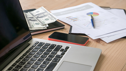 on the top of a desk, the picture presents a computer, a cellphone, multiple papers and money